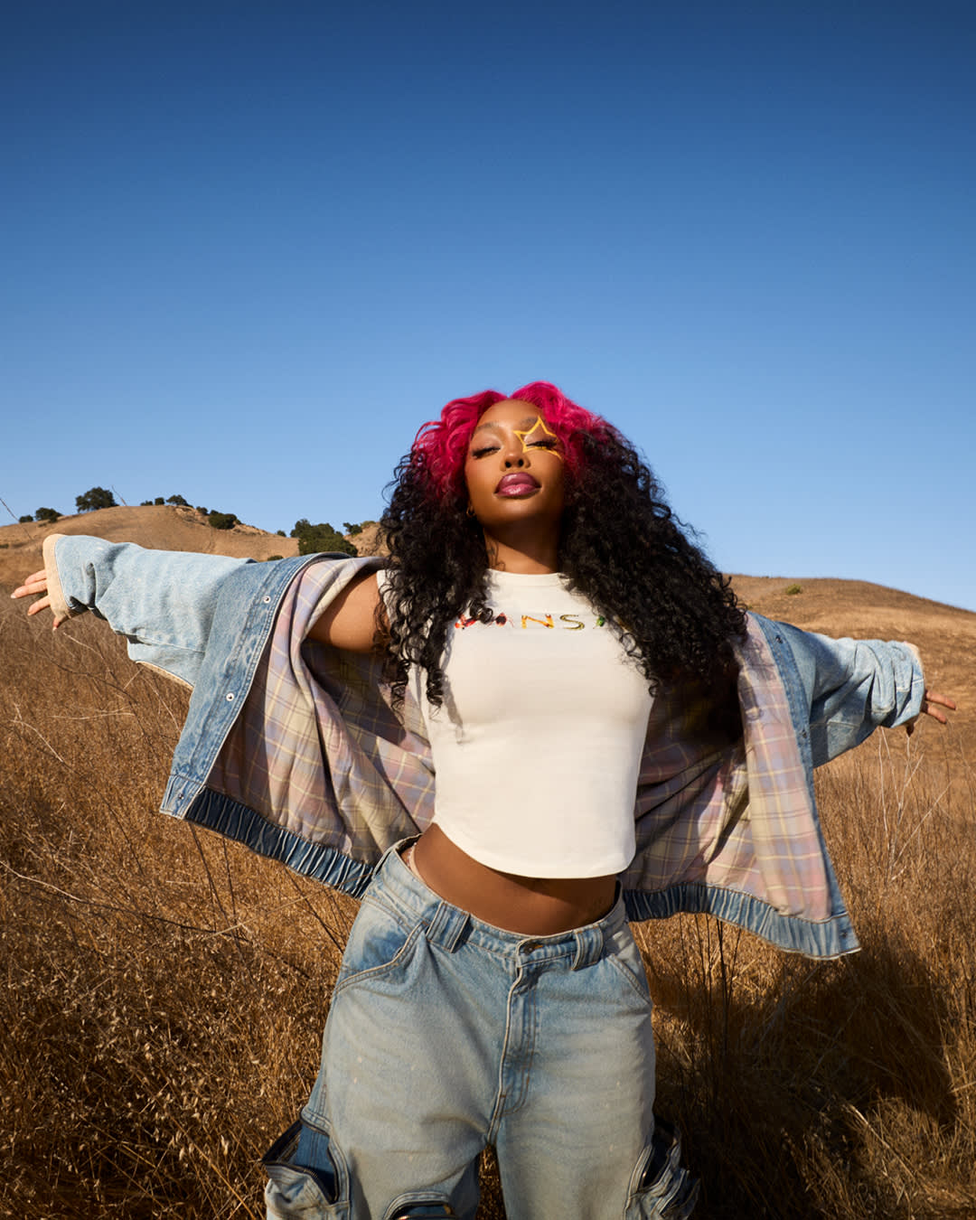SZA opens an oversized denim jacket in a sunlit California field, with pink-and-black hair and yellow star eye makeup